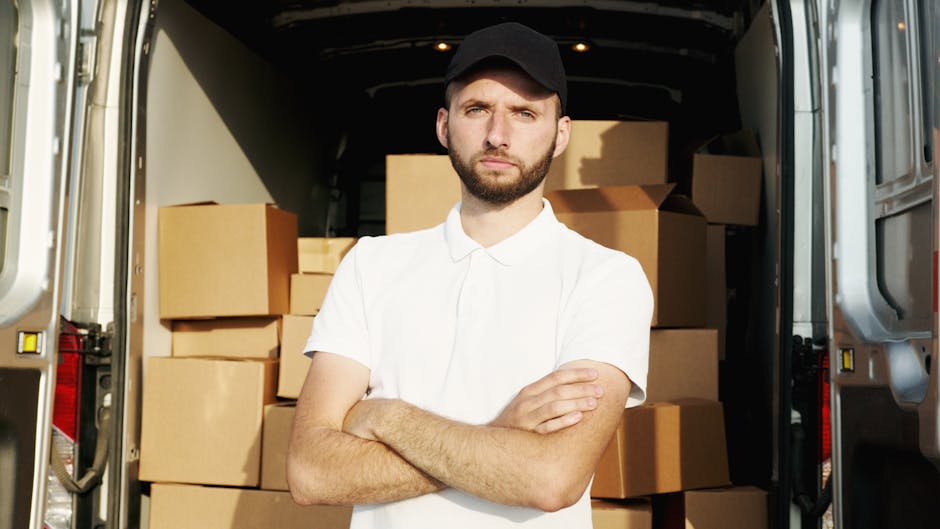 A young man wearing a white polo shirt and black cap stands inside the open rear of a delivery van filled with large cardboard boxes, some wrapped in plastic film, ready for home relocation. The van's interior is visible, showing a spacious cargo area with additional boxes stacked in the background, illustrating a furniture transport or packing and moving process. The man has arms crossed, with a neutral expression, standing on a paved surface outside a property, supporting professional removals services offered by Colliers Wood Man and Van as part of their house removals and relocation operations, with the scene emphasizing logistics, loading, and transport of household belongings.