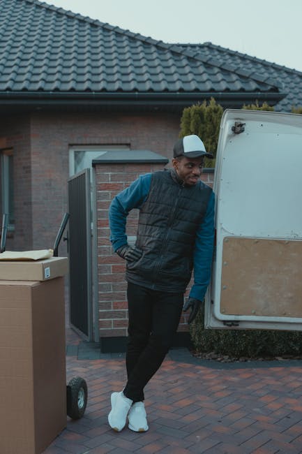 A man wearing a dark vest over a blue long-sleeve shirt, black pants, white sneakers, and a grey cap is standing beside an open rear door of a white moving van, which is parked on a paved driveway in front of a brick house with a tiled roof. The man is leaning slightly against the open door with one hand on his hip, observing the items being loaded or unloaded. In the foreground, there is a large cardboard box on a small hand trolley. The background features a hedge and a front garden, indicating a residential property. The scene appears to be part of a home relocation process involving furniture and packaging materials, with natural outdoor lighting and a clear sky. Colliers Wood Man and Van provides the moving service depicted in this image, illustrating the loading process of household items for an efficient move.