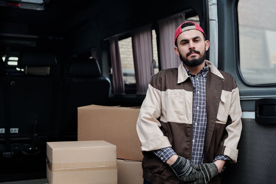 A young man wearing a white polo shirt and black cap stands inside the open rear of a delivery van filled with large cardboard boxes, some wrapped in plastic film, ready for home relocation. The van's interior is visible, showing a spacious cargo area with additional boxes stacked in the background, illustrating a furniture transport or packing and moving process. The man has arms crossed, with a neutral expression, standing on a paved surface outside a property, supporting professional removals services offered by Colliers Wood Man and Van as part of their house removals and relocation operations, with the scene emphasizing logistics, loading, and transport of household belongings.