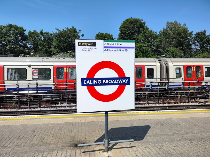 A clear view of a large white signage board at Ealing Broadway station, displaying directions for the way out, district line, Elizabeth line, and National Rail buses, with a focus on the red and white London Underground circle logo and the text 'EALING BROADWAY' in blue, positioned on a paved platform adjacent to a parked modern commuter train with red, white, and blue livery. The train's windows reveal interior seating, and the area includes a safety railing along the tracks with green trees and blue sky in the background, indicating a bright, sunny day, relevant to transport and home relocation logistics.