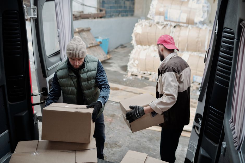 Two men engaged in a house removal process inside a storage or loading area, loading cardboard boxes into a large van. The man on the left, wearing a gray knit beanie, black gloves, and a quilted vest over a blue long-sleeve shirt, is lifting a medium-sized cardboard box. The man on the right, dressed in a beige and black work jacket, black gloves, and a red cap, is holding a smaller cardboard box and standing on the pavement just outside the van’s open rear doors. In the background, there are stacked large cardboard bundles wrapped with plastic, indicating packing materials, on a concrete surface, with some wooden pallets nearby. The scene is lit by natural daylight, suggesting a daytime moving or packing operation, typical of home relocation activities handled by Colliers Wood Man and Van as part of their removal services, including packing and furniture transport.