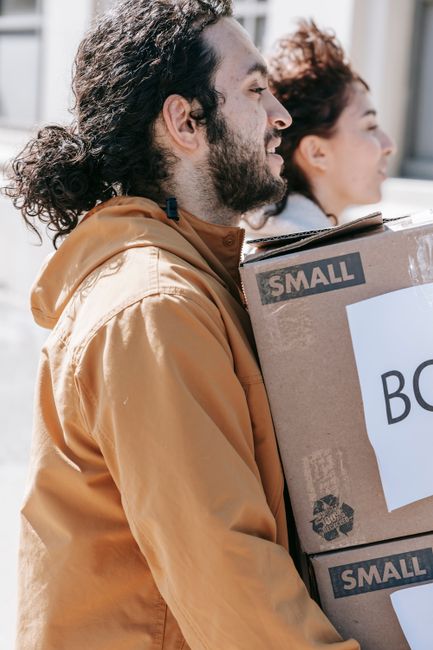 A man with dark curly hair and a beard, dressed in a tan jacket, is carrying a medium-sized cardboard box marked 'SMALL' with a white label attached. Behind him, a woman with reddish hair and light skin is visible, standing near a doorway or entrance of a property, possibly participating in a home relocation. The scene appears to be outdoors during daylight hours, with natural light illuminating the individuals. The box is part of a packing and moving process, indicative of furniture transport and loading activities associated with house removals. The background is slightly blurred, highlighting the focus on the man and the box, consistent with the context of professional removals, as offered by Colliers Wood Man and Van, on their page related to packing and load help for house moves.