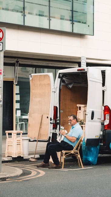 A man wearing glasses and a light blue shirt is seated on a wooden chair outside a white panel van, which is parked on the street near a modern building with large glass windows. His open van door rev