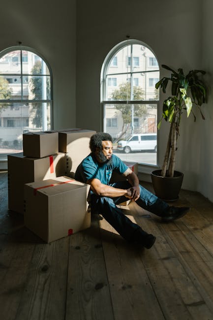 A man with curly black hair and dark skin, wearing a blue t-shirt and dark trousers, sits on the wooden floor of a room near two large arched windows that allow natural light to fill the space. Several cardboard boxes, some sealed with red tape, are stacked or placed on the floor next to him. The window reveals parked cars and a building exterior outside. A tall potted plant with broad green leaves is positioned in the corner of the room near the window. The room appears to be in the process of packing or moving, with the boxes likely prepared for transport as part of a home relocation. This scene is part of the loading and packing process during a house removal undertaken by Colliers Wood Man and Van, consistent with their same-day service in Colliers Wood, highlighting typical furniture transport and packing activities.
